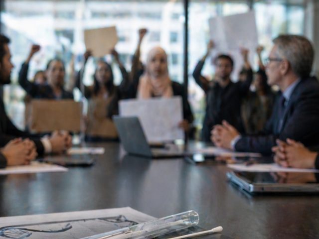 Boardroom executives meeting while a diverse group of employees protests with raised fists and signs outside the glass wall.