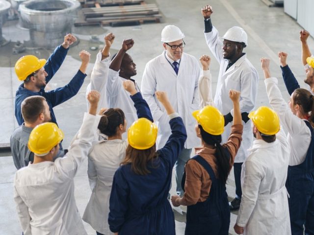 Group of factory workers and managers raising their fists together, illustrating union certification in a workplace.