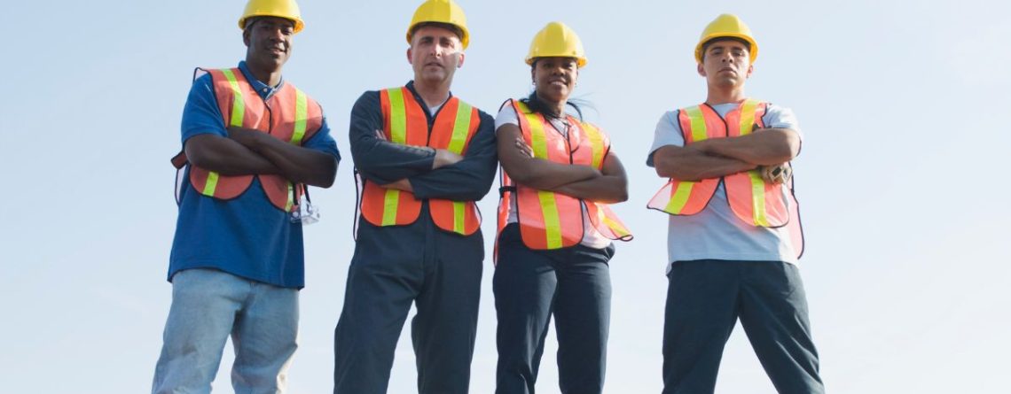 Four construction workers in hard hats and safety vests standing with arms crossed against a blue sky, illustrating workplace unionization.