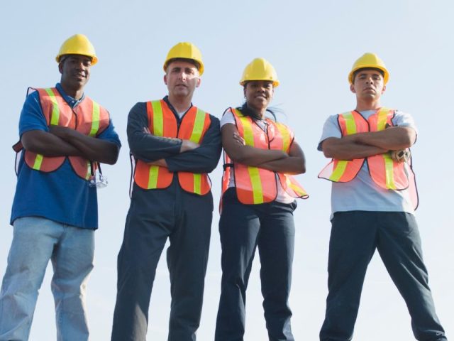 Four construction workers in hard hats and safety vests standing with arms crossed against a blue sky, illustrating workplace unionization.