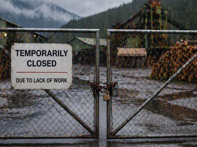 Padlocked chain-link gates at a BC lumber yard with a "Temporarily Closed Due to Lack of Work" sign, illustrating temporary layoffs.