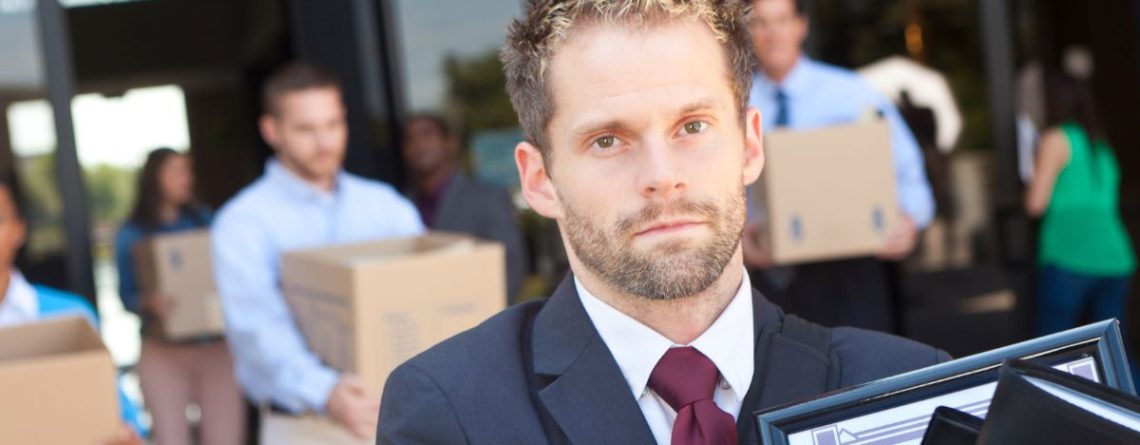 A man in close-up leaving an office building carrying a box of his desk belongings, with other laid-off employees doing the same in the background.