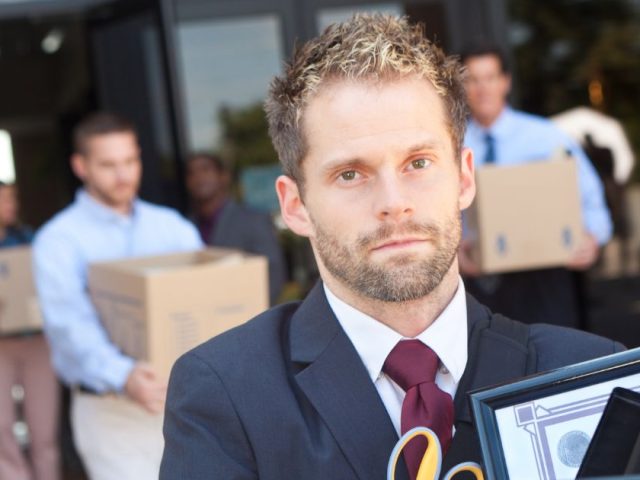 A man in close-up leaving an office building carrying a box of his desk belongings, with other laid-off employees doing the same in the background.