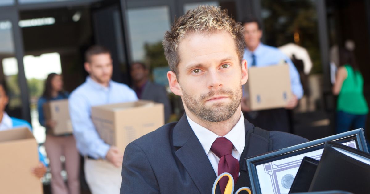 A man in close-up leaving an office building carrying a box of his desk belongings, with other laid-off employees doing the same in the background.